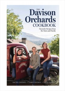 Cover image for “Davison Orchards Cookbook” by Rachel Davison, Tamra Davison, and Laura Shaw. The background image is a photograph of the authors, three smiling white women wearing jeans, sitting in an old, shiny, red pick-up truck with “Davison Orchards Est. 1933” scrawled on the door. Behind is a farm house and the Okanagan Valley further in the distance. In the top right corner is the title in a bold, dark blue, serif font it reads, “The Davison Orchards Cookbook: Favourite Recipes from the Farm and Family”. Along the bottom of the page is the three author names in a white, all-caps, serif font reading, “Rachel Davison, Tamra Davison, Laura Shaw”. The cover photo is by Callum Kyllonen and the cover design is by April Tran.